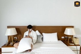 Bridegroom in Khao Yai National Park, Thailand, preparing for the wedding day with white cheek masks in a white, minimalistic hotel room setting on the bed.